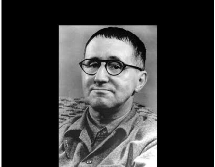 Black-and-white portrait of Bertolt Brecht — close-cropped hair, round wire-rim glasses, rough jacket, facing the camera with a slight half-smile.